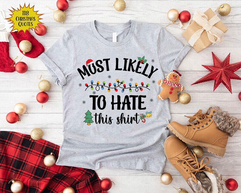 Może przedstawiać: Szary świąteczny T-shirt z napisem "Most Likely To Hate This Shirt" i łańcuchem światełek choinkowych. Koszulka ma świąteczny wz&oacute;r z małym drzewem choinkowym i dzwonkiem.