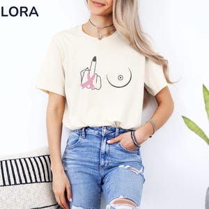 May include: Cream-colored t-shirt with a black graphic of a hand giving the middle finger, a breast, and a pink ribbon. The shirt has short sleeves and is paired with blue jeans. The brand name "MELORA SHIRTS" is visible in the top left corner.