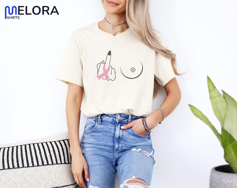 May include: Cream-colored t-shirt with a black graphic of a hand giving the middle finger, a breast, and a pink ribbon. The shirt has short sleeves and is paired with blue jeans. The brand name "MELORA SHIRTS" is visible in the top left corner.