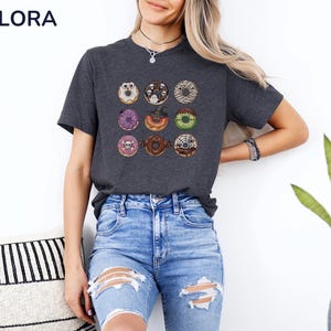 Puede incluir: Camiseta gris oscuro con una cuadr&iacute;cula de nueve donuts tem&aacute;ticos de Halloween. Cada donut tiene decoraciones &uacute;nicas, incluyendo fantasmas, calaveras y telara&ntilde;as. Los donuts son de varios colores, como morado, marr&oacute;n y verde.