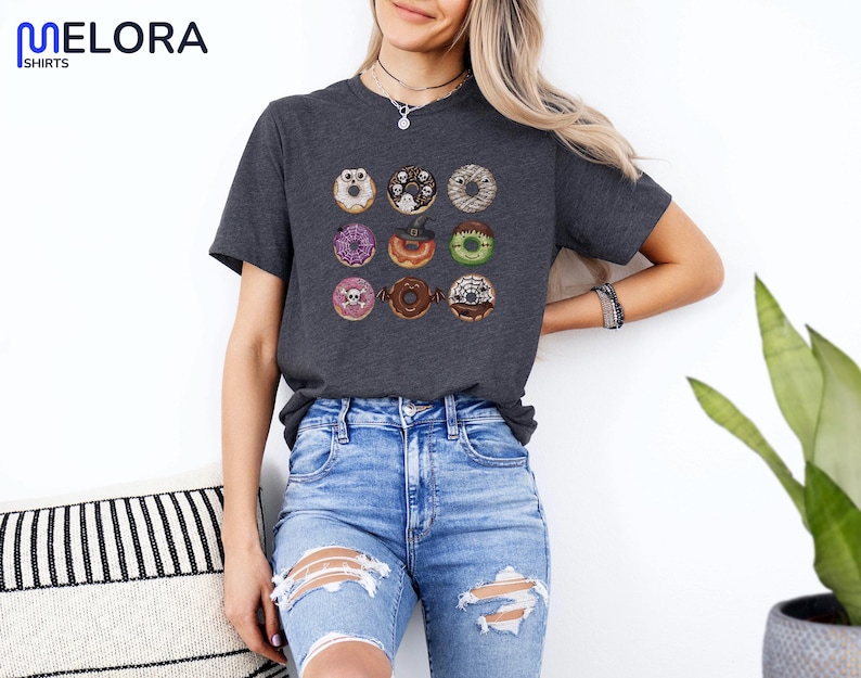 Puede incluir: Camiseta gris oscuro con una cuadr&iacute;cula de nueve donuts tem&aacute;ticos de Halloween. Cada donut tiene decoraciones &uacute;nicas, incluyendo fantasmas, calaveras y telara&ntilde;as. Los donuts son de varios colores, como morado, marr&oacute;n y verde.