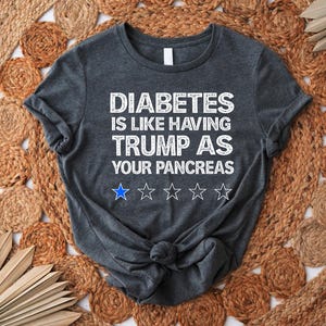 Puede incluir: Una camiseta gris oscuro con el texto "DIABETES IS LIKE HAVING TRUMP AS YOUR PANCREAS" en blanco. El texto est&aacute; en una fuente desgastada y hay cinco estrellas blancas debajo del texto.