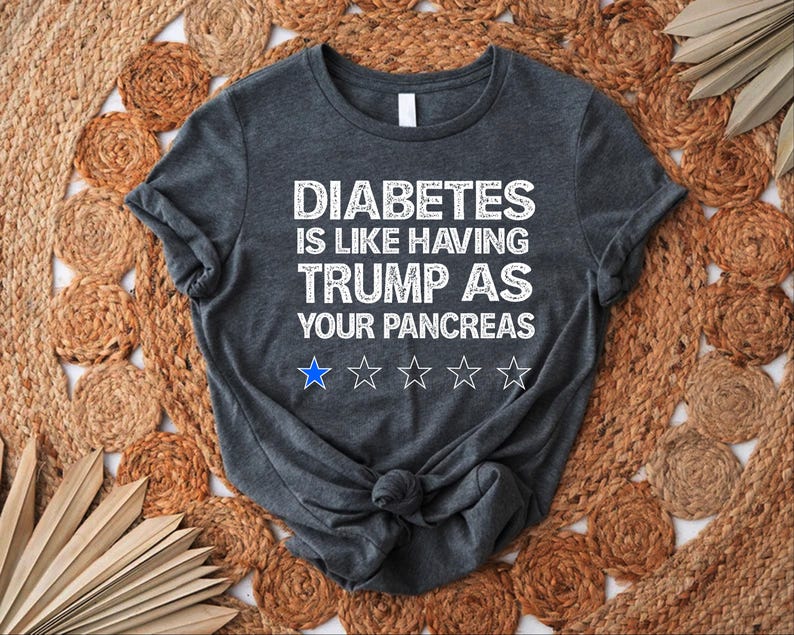 Puede incluir: Una camiseta gris oscuro con el texto "DIABETES IS LIKE HAVING TRUMP AS YOUR PANCREAS" en blanco. El texto est&aacute; en una fuente desgastada y hay cinco estrellas blancas debajo del texto.