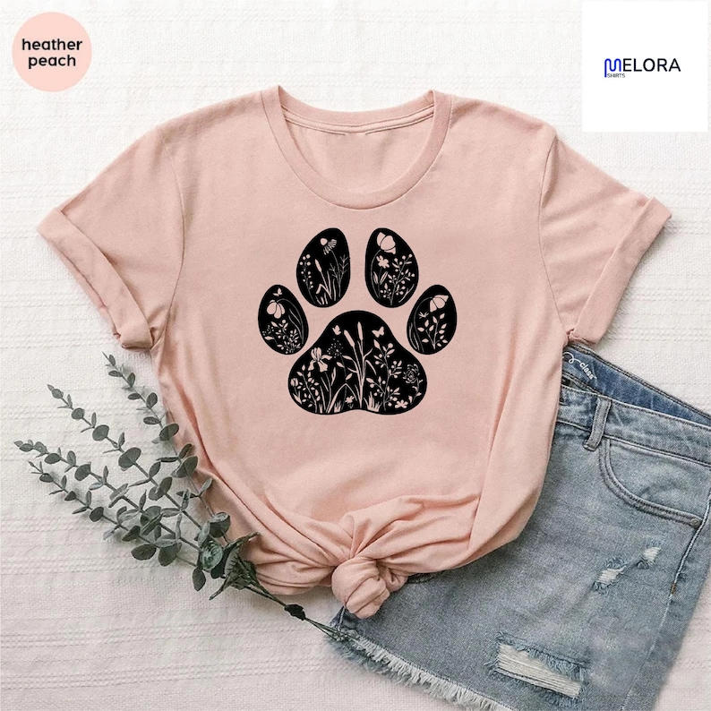 May include: A heather peach t-shirt featuring a black paw print design filled with floral and butterfly silhouettes. The shirt has a crew neck and short sleeves. The text "heather peach" is in a circle on the upper left.
