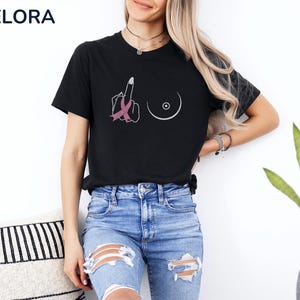 May include: Black t-shirt with a graphic design featuring a hand giving the middle finger with a pink ribbon, and a breast outline. The shirt has the word "MELORA SHIRTS" in the upper left corner.