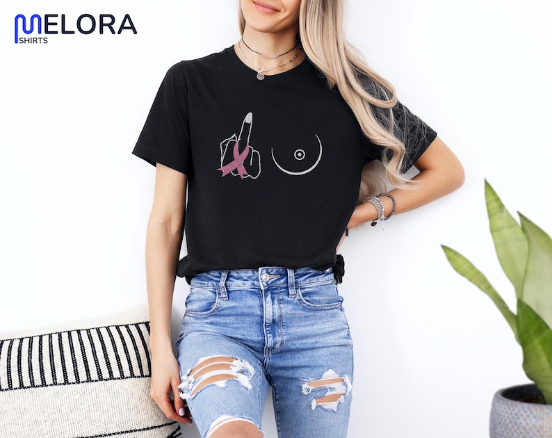 May include: Black t-shirt with a graphic design featuring a hand giving the middle finger with a pink ribbon, and a breast outline. The shirt has the word "MELORA SHIRTS" in the upper left corner.
