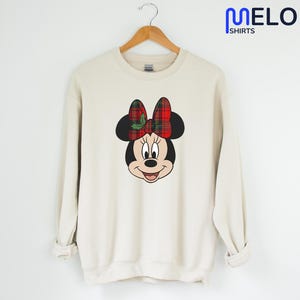 May include: A cream-coloured sweatshirt with a Minnie Mouse graphic. The graphic depicts Minnie's face with a red and green plaid bow. The sweatshirt hangs on a wooden hanger. The text "MELORA SHIRTS" is in the upper right corner.