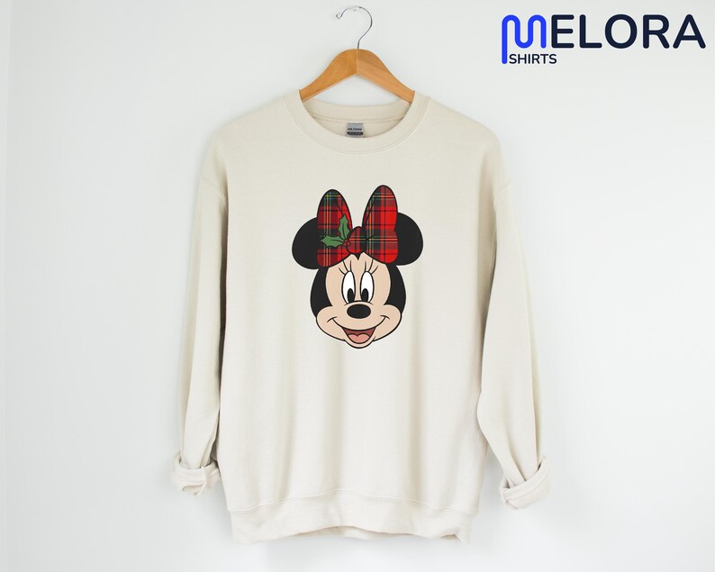 May include: A cream-coloured sweatshirt with a Minnie Mouse graphic. The graphic depicts Minnie's face with a red and green plaid bow. The sweatshirt hangs on a wooden hanger. The text "MELORA SHIRTS" is in the upper right corner.