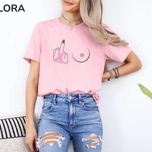 May include: Pink t-shirt with a graphic design featuring a hand giving the middle finger, a breast, and a pink ribbon. The shirt has a crew neck and short sleeves. The brand name "MELORA SHIRTS" is printed in blue on the upper left.