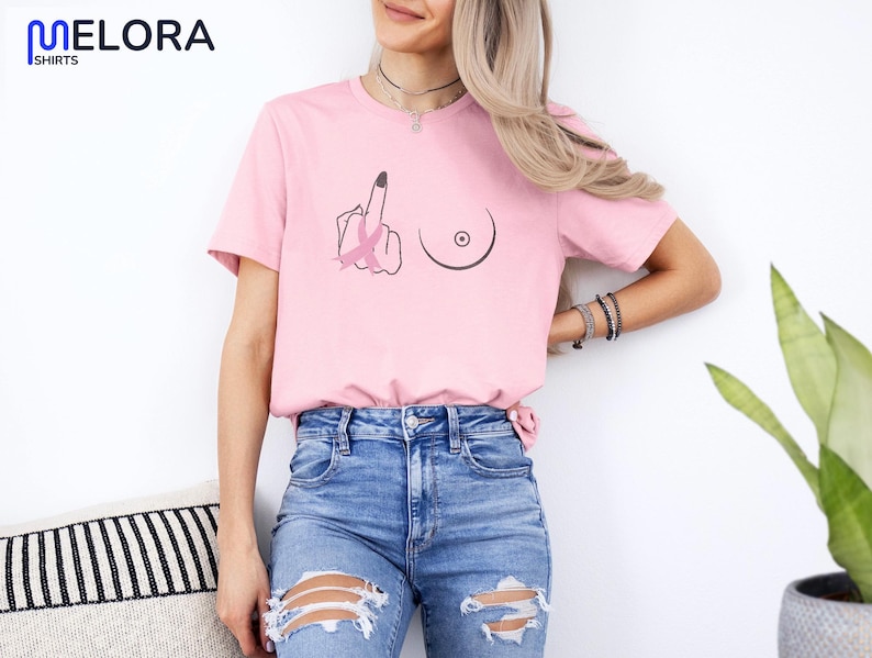 May include: Pink t-shirt with a graphic design featuring a hand giving the middle finger, a breast, and a pink ribbon. The shirt has a crew neck and short sleeves. The brand name "MELORA SHIRTS" is printed in blue on the upper left.