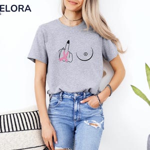 May include: A heather gray t-shirt with a graphic design featuring a hand giving the middle finger, a breast, and a pink ribbon. The shirt has short sleeves and a crew neck.