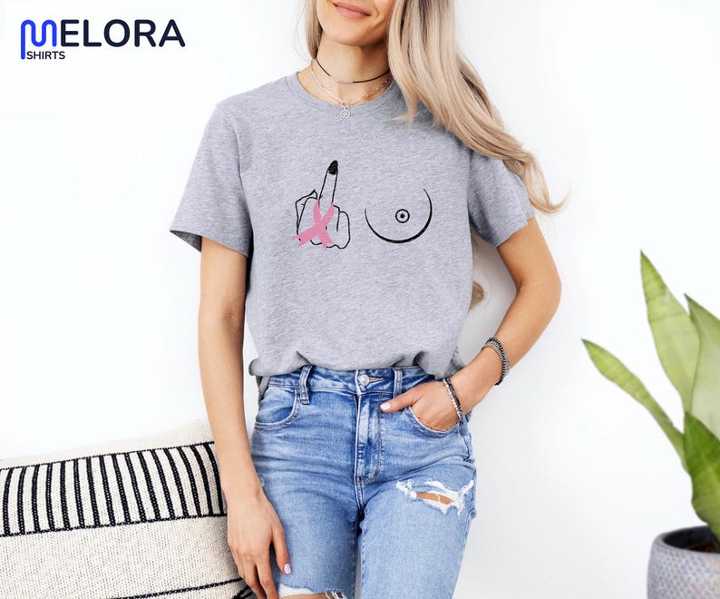 May include: A heather gray t-shirt with a graphic design featuring a hand giving the middle finger, a breast, and a pink ribbon. The shirt has short sleeves and a crew neck.