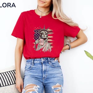 K&ouml;nnte beinhalten: Rotes T-Shirt mit einer Grafik der Freiheitsstatue, die ein Schild mit der Aufschrift "IS HE DEAD YET?" und einem amerikanischen Flaggen-Design h&auml;lt. Das Shirt ist ein kurz&auml;rmeliges Rundhals-Shirt.