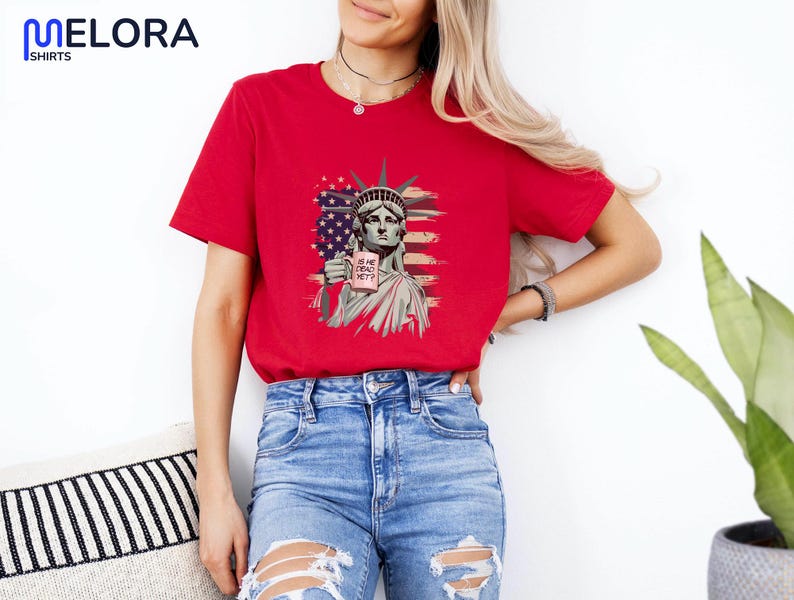 K&ouml;nnte beinhalten: Rotes T-Shirt mit einer Grafik der Freiheitsstatue, die ein Schild mit der Aufschrift "IS HE DEAD YET?" und einem amerikanischen Flaggen-Design h&auml;lt. Das Shirt ist ein kurz&auml;rmeliges Rundhals-Shirt.