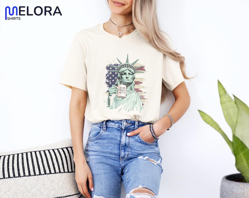 K&ouml;nnte beinhalten: Cremefarbenes T-Shirt mit einer Grafik der Freiheitsstatue, die ein Schild mit der Aufschrift "IS HE DEAD YET?" h&auml;lt. Das Design beinhaltet einen Hintergrund der amerikanischen Flagge. Das Shirt wird mit blauen Jeans getragen.