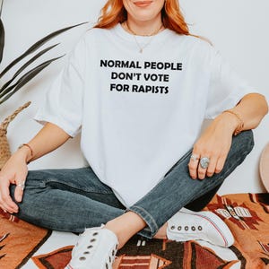 May include: White t-shirt with the text "NORMAL PEOPLE DON'T VOTE FOR RAPISTS" in black block letters. The person is wearing blue jeans and white trainers. The person is sitting on a patterned blanket.