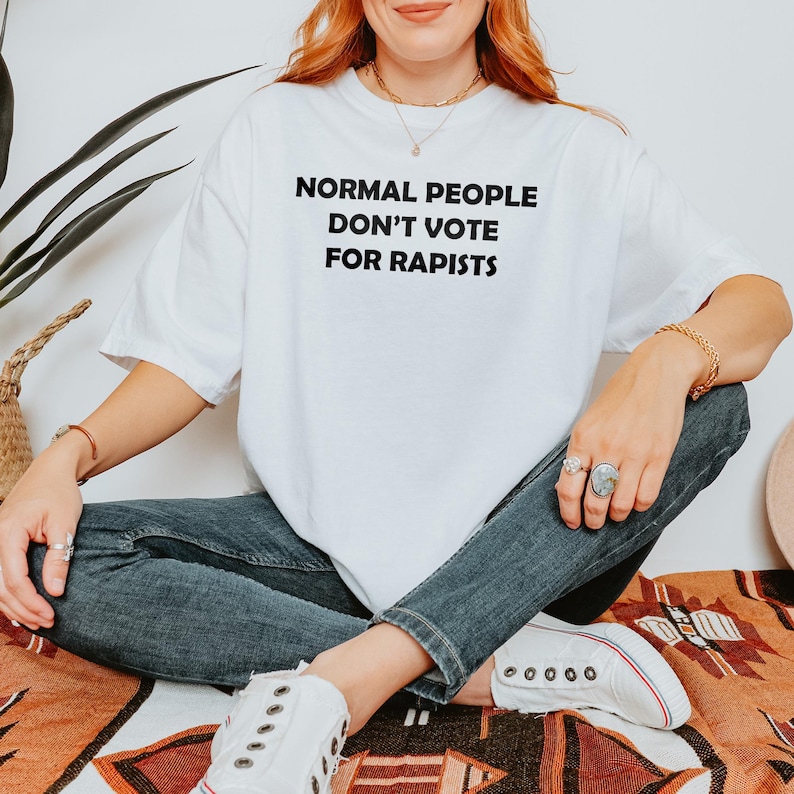 May include: White t-shirt with the text "NORMAL PEOPLE DON'T VOTE FOR RAPISTS" in black block letters. The person is wearing blue jeans and white trainers. The person is sitting on a patterned blanket.