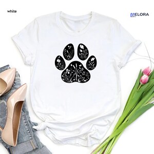 May include: White t-shirt featuring a black paw print design filled with floral and botanical illustrations. The shirt has short sleeves and a crew neck. The word "white" is in the top left corner.