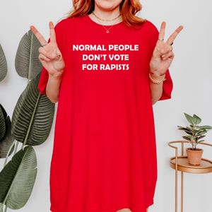 May include: A vibrant red t-shirt featuring the white text "NORMAL PEOPLE DON'T VOTE FOR RAPISTS". The shirt is worn by a person making a peace sign with their fingers.