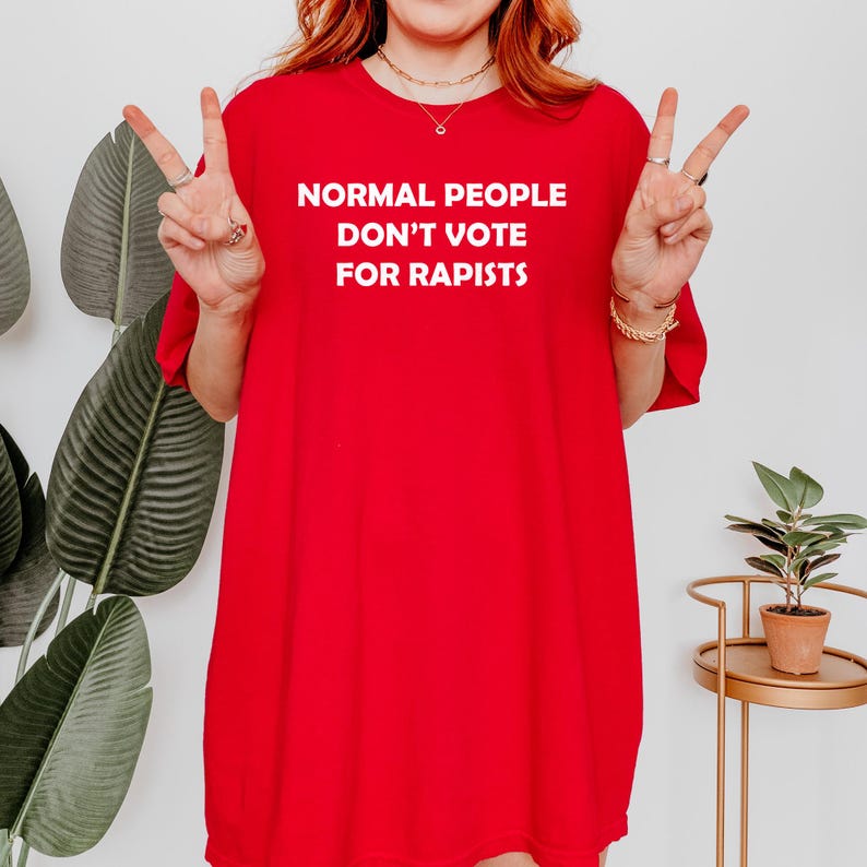 May include: A vibrant red t-shirt featuring the white text "NORMAL PEOPLE DON'T VOTE FOR RAPISTS". The shirt is worn by a person making a peace sign with their fingers.