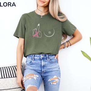 May include: Olive green t-shirt with a graphic design featuring a hand giving the middle finger with a pink ribbon, and a breast outline. The shirt has the word "MELORA SHIRTS" at the top left.