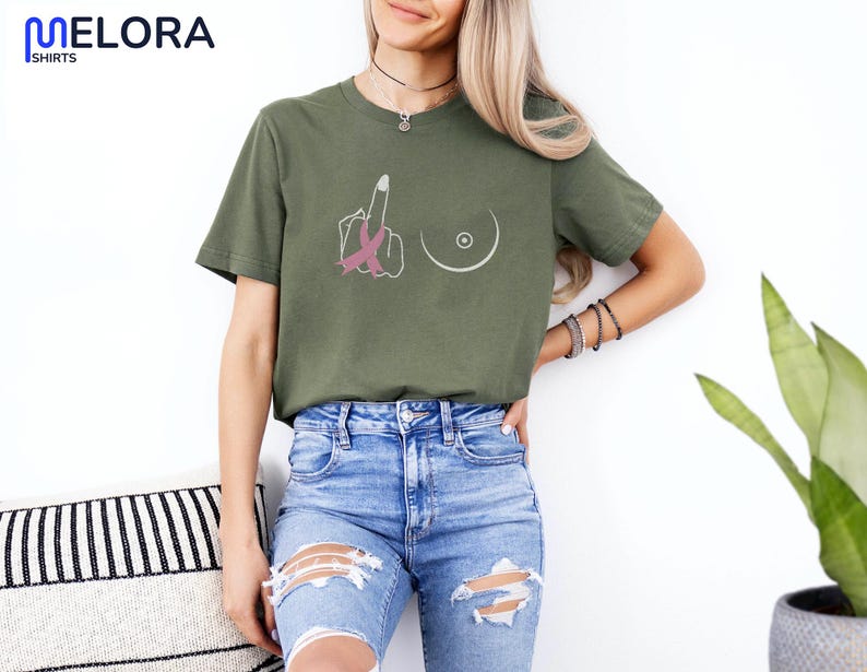 May include: Olive green t-shirt with a graphic design featuring a hand giving the middle finger with a pink ribbon, and a breast outline. The shirt has the word "MELORA SHIRTS" at the top left.
