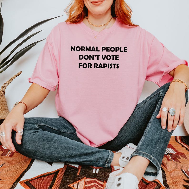 May include: A pale pink t-shirt with the slogan "NORMAL PEOPLE DON'T VOTE FOR RAPISTS" in black lettering. The shirt is paired with blue denim jeans and white trainers. The person is sitting on a patterned blanket.