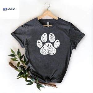 May include: Dark gray t-shirt featuring a white paw print design filled with floral and botanical illustrations. The shirt is displayed on a wooden hanger. The brand name "MELORA SPORTS" is visible in the upper left corner.