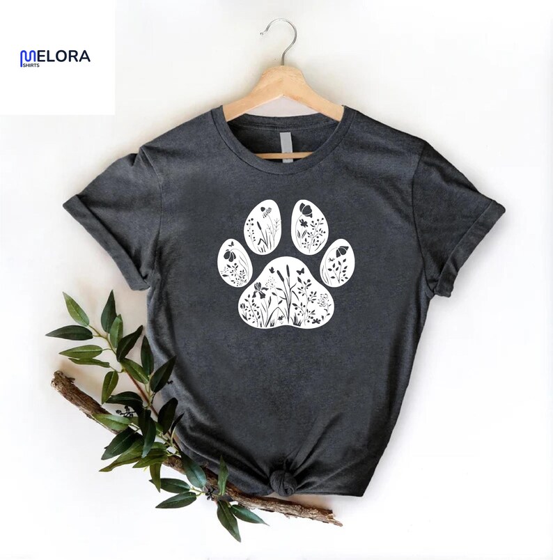 May include: Dark gray t-shirt featuring a white paw print design filled with floral and botanical illustrations. The shirt is displayed on a wooden hanger. The brand name "MELORA SPORTS" is visible in the upper left corner.