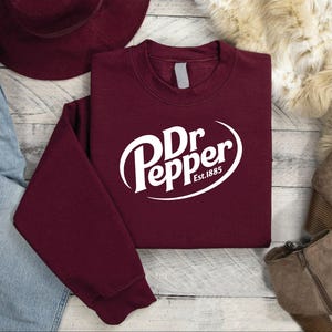 May include: A maroon sweatshirt with the Dr Pepper logo in white. The logo includes the brand name and "Est.1885" in a white oval. The sweatshirt is folded on a light wood surface.