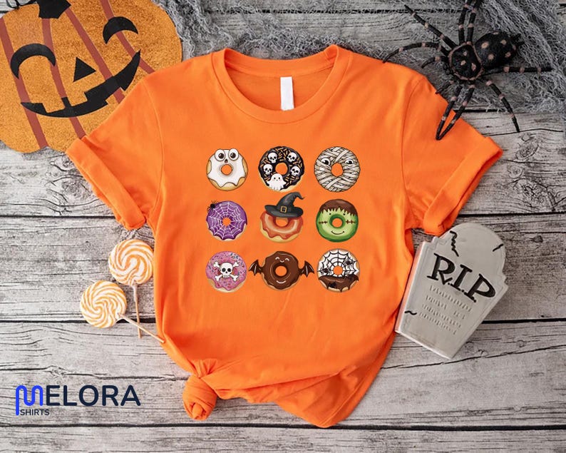 Puede incluir: Camiseta naranja brillante con un dise&ntilde;o de donuts de Halloween. Los donuts presentan temas espeluznantes como momias, brujas y ara&ntilde;as. La camiseta es de Melora Shirts.