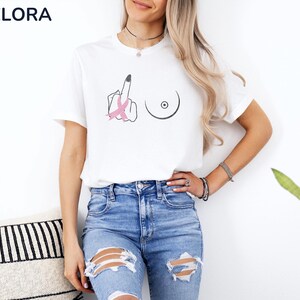 May include: White t-shirt with a graphic design featuring a middle finger with a pink ribbon and a breast outline. The shirt is paired with blue jeans. The shirt is from Melora Shirts.