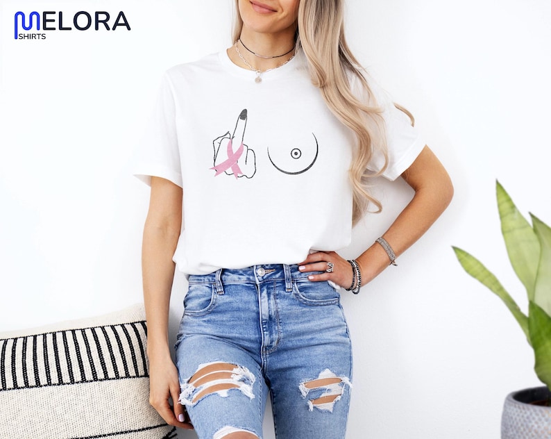 May include: White t-shirt with a graphic design featuring a middle finger with a pink ribbon and a breast outline. The shirt is paired with blue jeans. The shirt is from Melora Shirts.