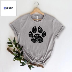 May include: A gray t-shirt featuring a black paw print design filled with floral illustrations. The shirt is displayed on a wooden hanger. The brand name "MELORA SHIRTS" is visible in the upper left corner.