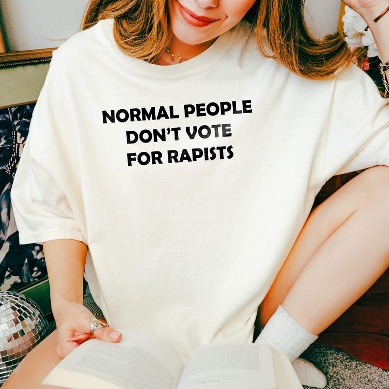 May include: A cream-coloured t-shirt featuring the black text "NORMAL PEOPLE DON'T VOTE FOR RAPISTS". The person wearing the shirt is holding a book. The image suggests a relaxed, everyday style.