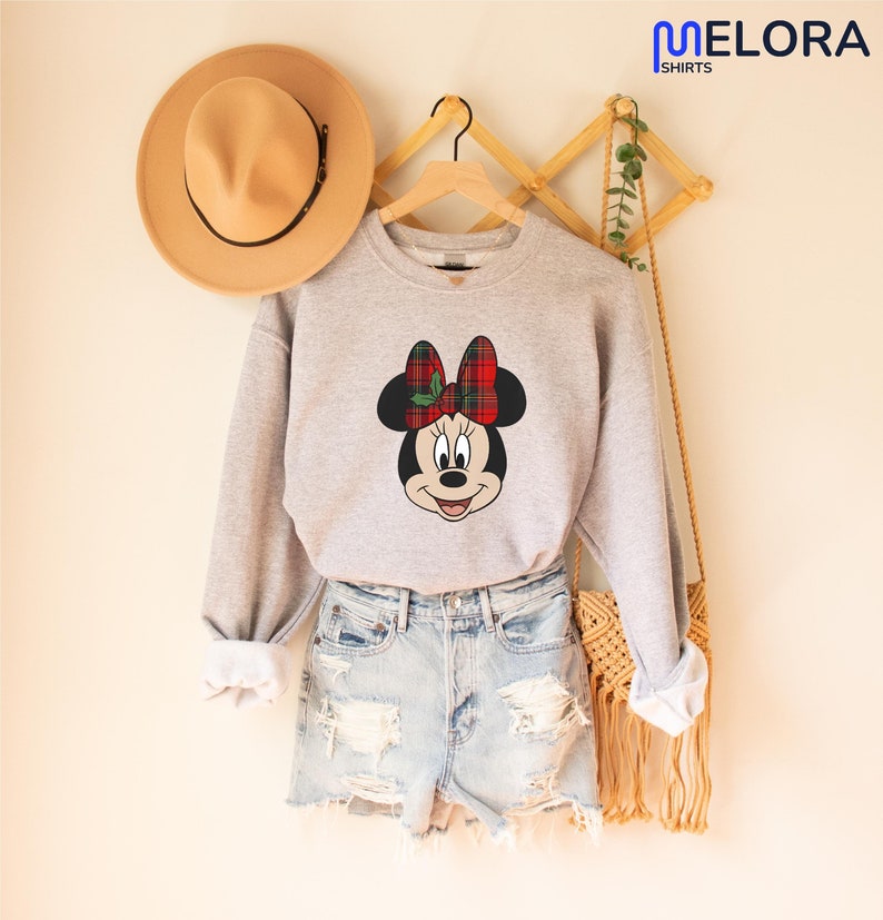 May include: A light grey sweatshirt with a Minnie Mouse graphic, sporting a red and green tartan bow. The sweatshirt is displayed with denim shorts and a tan hat. The image is from Melora Shirts.