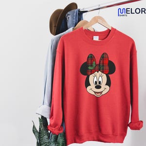 May include: Red sweatshirt with a Minnie Mouse graphic, a plaid bow and holly. The sweatshirt is hanging on a clothes rail with other items. The brand name "MELORA SHIRTS" is visible in the upper right corner.