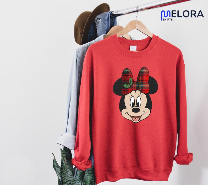 May include: Red sweatshirt with a Minnie Mouse graphic, a plaid bow and holly. The sweatshirt is hanging on a clothes rail with other items. The brand name "MELORA SHIRTS" is visible in the upper right corner.