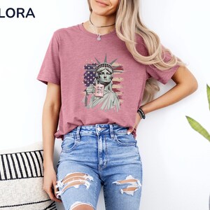 K&ouml;nnte beinhalten: Ein mauvefarbenes T-Shirt mit einer Grafik der Freiheitsstatue, die ein Schild mit der Aufschrift "IS SHE DEAD YET?" h&auml;lt. Das Design enth&auml;lt ein amerikanisches Flaggenmotiv. Das Shirt ist Teil der Melora Shirts Kollektion.