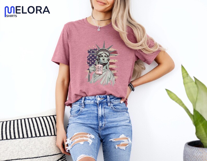 K&ouml;nnte beinhalten: Ein mauvefarbenes T-Shirt mit einer Grafik der Freiheitsstatue, die ein Schild mit der Aufschrift "IS SHE DEAD YET?" h&auml;lt. Das Design enth&auml;lt ein amerikanisches Flaggenmotiv. Das Shirt ist Teil der Melora Shirts Kollektion.