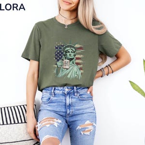 K&ouml;nnte beinhalten: Olivgr&uuml;nes T-Shirt mit einer Grafik der Freiheitsstatue, die ein Schild mit der Aufschrift "IS HE DEAD YET?" vor dem Hintergrund der amerikanischen Flagge h&auml;lt. Das T-Shirt hat kurze &Auml;rmel und einen Rundhalsausschnitt.