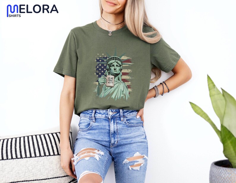 K&ouml;nnte beinhalten: Olivgr&uuml;nes T-Shirt mit einer Grafik der Freiheitsstatue, die ein Schild mit der Aufschrift "IS HE DEAD YET?" vor dem Hintergrund der amerikanischen Flagge h&auml;lt. Das T-Shirt hat kurze &Auml;rmel und einen Rundhalsausschnitt.