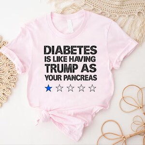 Puede incluir: Camiseta rosa con texto negro que dice "La diabetes es como tener a Trump como p&aacute;ncreas". Hay cinco estrellas negras debajo del texto, con una estrella azul en el medio.