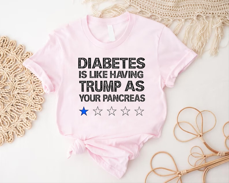 Puede incluir: Camiseta rosa con texto negro que dice "La diabetes es como tener a Trump como p&aacute;ncreas". Hay cinco estrellas negras debajo del texto, con una estrella azul en el medio.