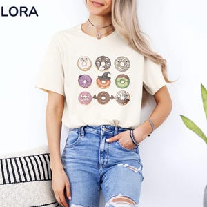 Puede incluir: Camiseta color crema con una cuadr&iacute;cula de nueve ilustraciones de donuts con tem&aacute;tica de Halloween. Los donuts tienen varios toppings y decoraciones, incluyendo calaveras, sombreros de bruja y vendas de momia. La camiseta se combina con vaqueros azules.
