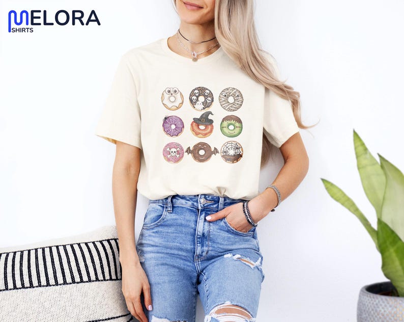 Puede incluir: Camiseta color crema con una cuadr&iacute;cula de nueve ilustraciones de donuts con tem&aacute;tica de Halloween. Los donuts tienen varios toppings y decoraciones, incluyendo calaveras, sombreros de bruja y vendas de momia. La camiseta se combina con vaqueros azules.