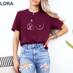 May include: Burgundy t-shirt with a graphic design featuring a middle finger with a pink ribbon and a breast outline. The shirt is paired with blue jeans. The brand name "MELORA SHIRTS" is visible in the top left corner.