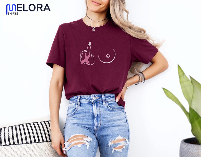 May include: Burgundy t-shirt with a graphic design featuring a middle finger with a pink ribbon and a breast outline. The shirt is paired with blue jeans. The brand name "MELORA SHIRTS" is visible in the top left corner.