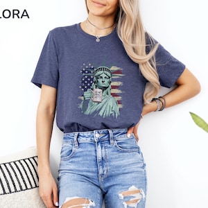 K&ouml;nnte beinhalten: Marineblaues T-Shirt mit einer Grafik der Freiheitsstatue, die einen Becher mit dem Text "IS SHE DEAD YET?" vor einer verblassten amerikanischen Flagge h&auml;lt. Das Shirt hat einen Rundhalsausschnitt und ist aus weichem Material.