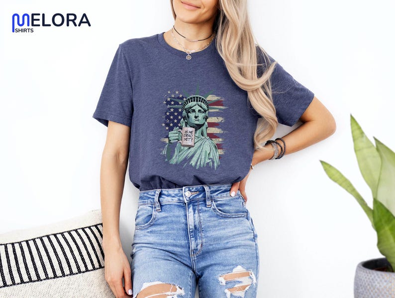 K&ouml;nnte beinhalten: Marineblaues T-Shirt mit einer Grafik der Freiheitsstatue, die einen Becher mit dem Text "IS SHE DEAD YET?" vor einer verblassten amerikanischen Flagge h&auml;lt. Das Shirt hat einen Rundhalsausschnitt und ist aus weichem Material.